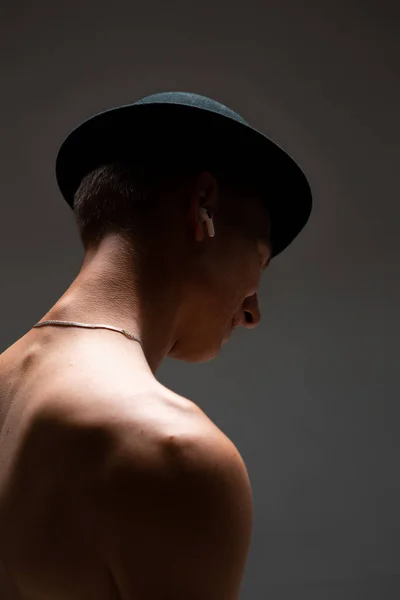 Joven chico fresco en sombrero negro y en un auricular inalámbrico escuchar la canción sin camisa sobre fondo negro. Cartel musical —  Fotos de Stock