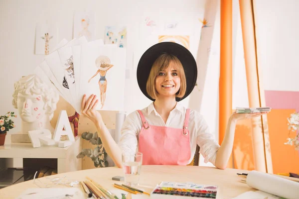 Kreativ glad flicka i hatt artist eller illustratör sitter med pengar och teckningar i sin verkstad — Stockfoto