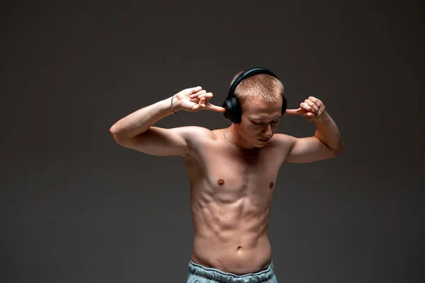 一个穿着带裸体躯干的无线耳机的很酷的年轻人，在黑背景音乐工作室里听音乐吧 — 图库照片