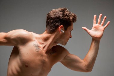 Kablosuz kulaklıklı havalı seksi adam. Gri arka planda çalışan çıplak gövdeli şarkı dinliyor. Dövmeli vücut.