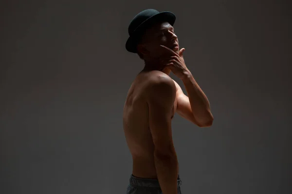 Snygg ung kille skådespelare i hatt står utan skjorta i studio isolerad på svart bakgrund — Stockfoto