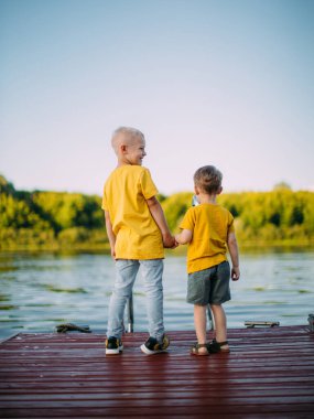 Havalı, gülümseyen çocuk ve kardeşi el ele tutuşmuş ve rıhtımda durmuş nehri izliyorlar. Arka plan