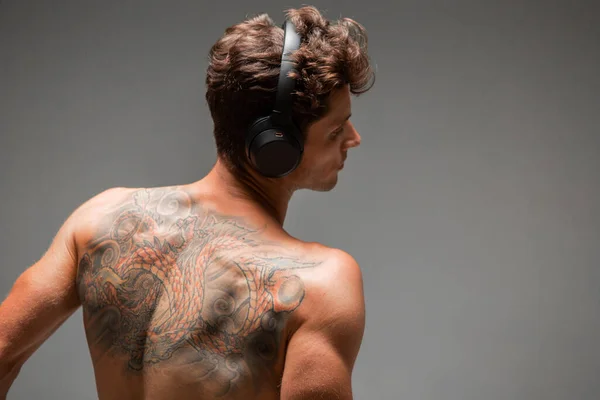 Menő fiatal srác vezeték nélküli fejhallgatóval hallgat dalt meztelen háttal elszigetelt szürke háttérrel. Tetovált test — Stock Fotó