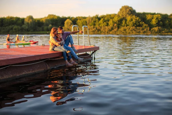 Fiatal család ül a mólón a folyón vagy a tavon nyáron. Fényképezés reklám vagy blog a családról és az utazásról — Stock Fotó