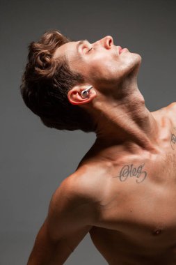 Kablosuz kulaklıklı, çıplak vücutlu ve gözleri gri arka planda olan havalı bir genç. Müzik posteri. Dövmeli vücut.