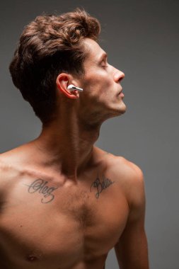 Kablosuz kulaklıklı havalı genç adam gri arka planda tişörtsüz şarkı dinliyor. Müzik posteri. Dövmeli vücut..