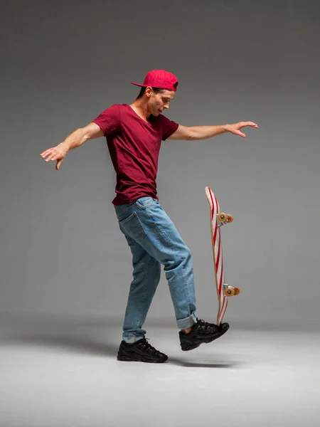 Cool guy skateboarder keeps skateboard on leg in studio on grey background. Photography about skateboarding and balance