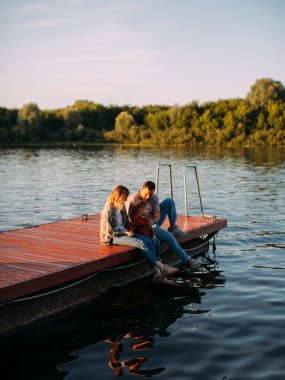 Genç bir aile yazın nehir veya gölde iskelede oturuyor. Aile ve seyahat hakkında reklam veya blog için fotoğraf