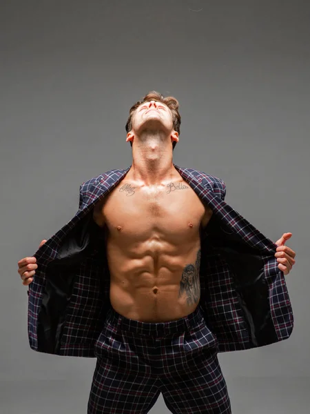 Stilig ung man i en vidöppen jacka med naken bål står i studio på grå bakgrund. Kropp med tatuering — Stockfoto