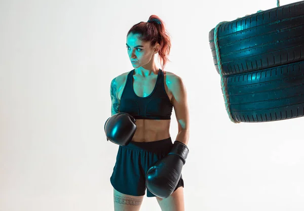 Atlético lutador menina em luvas de boxe posando em luz de néon com saco de perfuração de pneus. Cartaz de artes marciais mistas — Fotografia de Stock