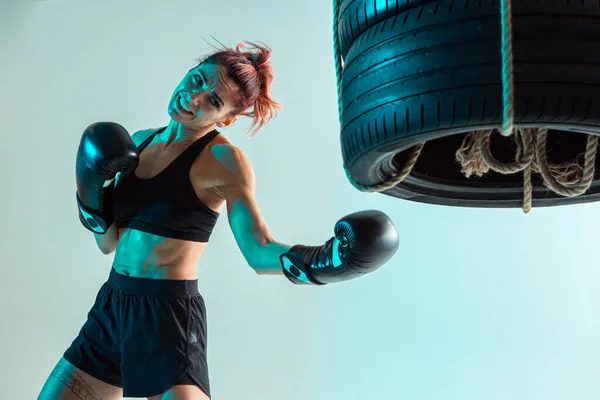 Athletic female fighter trains uppercut on punching bag made of tires in studio in neon light. Mixed martial arts poster