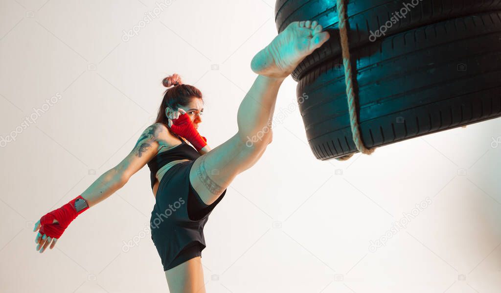 Cool girl fighter practicing kicks using tires in studio isolated on white background in neon. Mixed martial arts poster