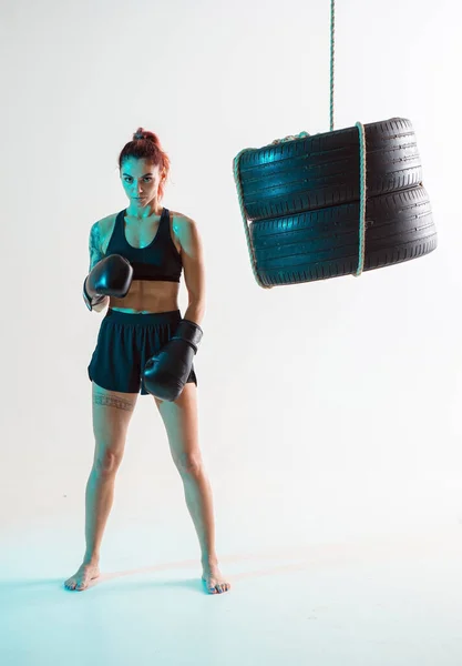 Boxer female fighter in boxing gloves posing in confident stance near punching bag of tires. Mixed martial arts poster