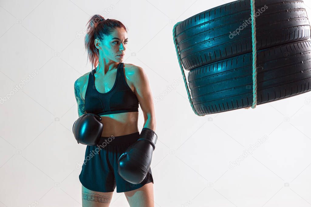Athletic girl fighter in boxing gloves posing in neon light with punching bag made of tires. Mixed martial arts poster