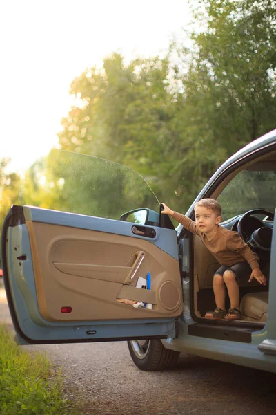 Χαμογελαστό αγοράκι, μικρός οδηγός χούλιγκαν, κάθεται στο αυτοκίνητο. Καλοκαιρινή φωτογραφία για διαφήμιση ή blog σχετικά με το οικογενειακό ταξίδι — Φωτογραφία Αρχείου