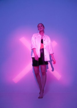 Havalı genç bayan model Violet stüdyosunun arka planında pembe haçla neon ışıkta duruyor.
