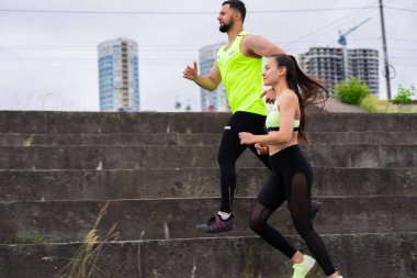 Şık spor kıyafetleri içinde atletik erkek ve kız şehirde merdivenlerde koşuyor, blog için fotoğraf ve sağlıklı yaşam tarzı reklamı çekiyor.