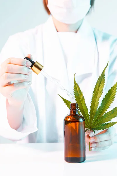 Gros plan du médecin en masque et peignoir avec une feuille de chanvre, une pipette et une bouteille fictive de produit à base d'huile de cannabis — Photo