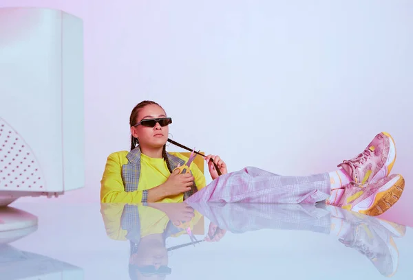 Stijlvolle vrouwelijke kantoormedewerker in een zonnebril zittend met verouderde computer met voeten op tafel. Gekleurd neonlicht — Stockfoto