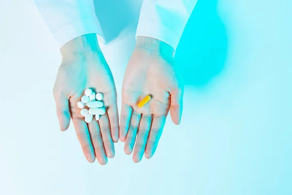 Куча белых таблеток и одна желтая капсула в руках человека в синем неоновом свете, вид сверху. Альтернативная медицина — стоковое фото