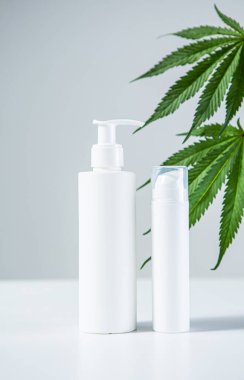 Bir çift beyaz CBD doğal ürünleri ve gri stüdyo arka planında kenevir yaprakları olan boş şişeleri taklit eder..