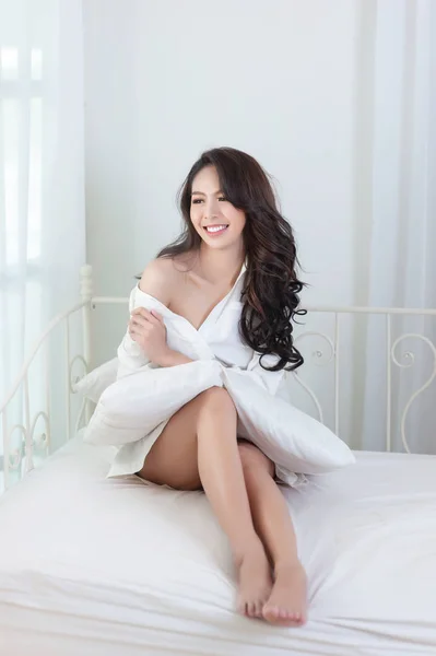 Seksi beyaz bir gömlek güzel bir Asyalı kadın mutlu bir beyaz yatakta gülümsüyor 