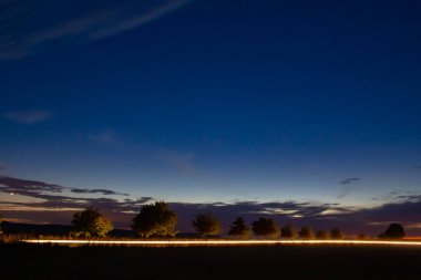 Kırsal bir yolda araba süren ve gün batımında ağaçları aydınlatan bir arabanın farları.