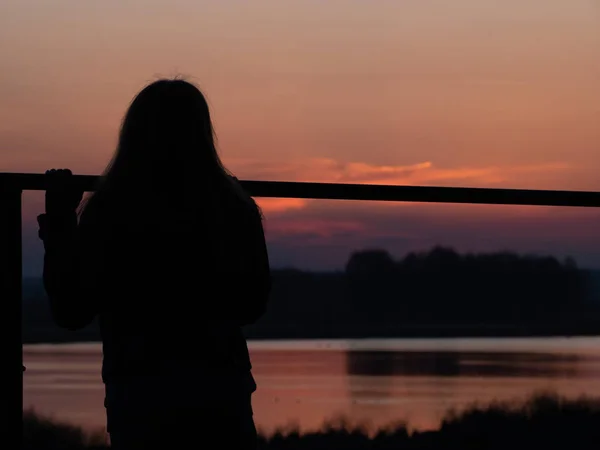 siluet bir genç kızın alacakaranlıkta günbatımı holding çit, uzun saçlı. güzel kırsal manzara manzara manzara nehir ya da göl üzerinde arka plan ile. düşük anahtar fotoğraf