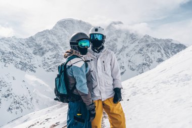 Kar kayakçıları kız ve erkek, arka planda gururla, kameraya bakıyor, dağları yansıtan mavi snowboard gözlükleri takıyor.