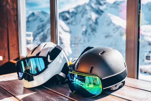 two ski helmets with ski goggles lie on the table, in the background a view of the mountains through the window