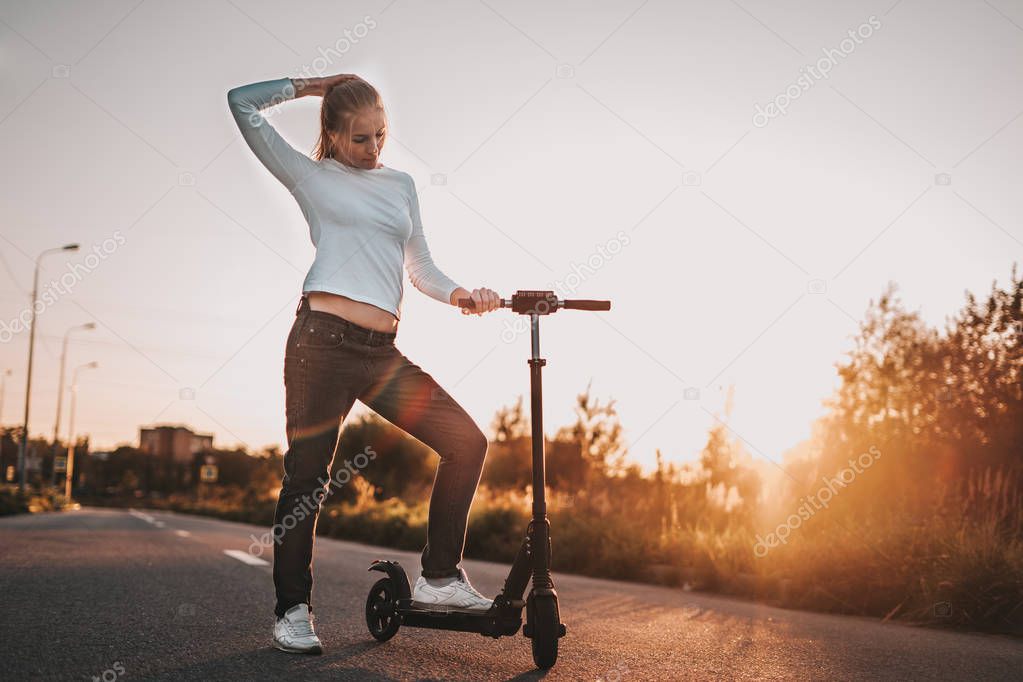 Young beautiful girl riding an electric scooter in the summer on the street, against the sunset, there is free space for advertising against the sky