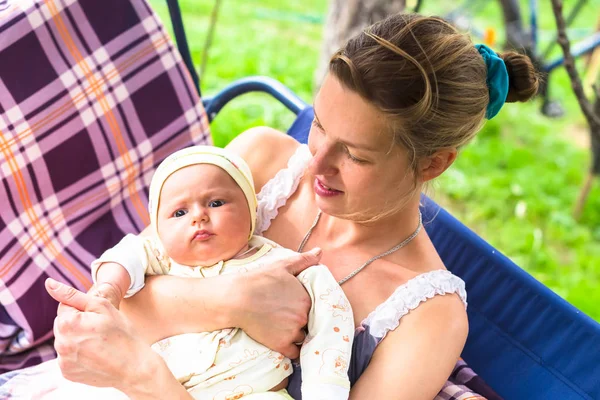 Anne ve bebek bahçede yaz bir salıncak üzerinde durmak. Kalıcı makyaj, doğal ve tedavi olmadan anne. Bebek üç aylık küçük bir kız gibi. Çocuk yawns.