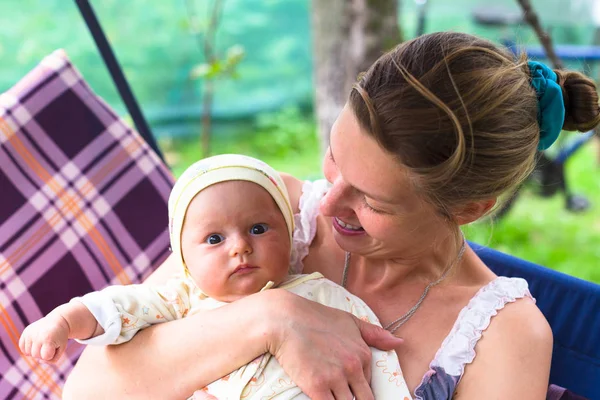 Anne ve bebek bahçede yaz bir salıncak üzerinde durmak. Kalıcı makyaj, doğal ve tedavi olmadan anne. Bebek üç aylık küçük bir kız gibi. Çocuk yawns.