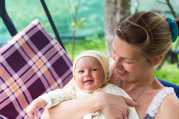Anne ve bebek bahçede yaz bir salıncak üzerinde durmak. Kalıcı makyaj, doğal ve tedavi olmadan anne. Bebek üç aylık küçük bir kız gibi. Çocuk yawns.