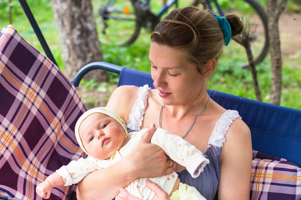 Anne ve bebek bahçede yaz bir salıncak üzerinde durmak. Kalıcı makyaj, doğal ve tedavi olmadan anne. Bebek üç aylık küçük bir kız gibi. Çocuk yawns.