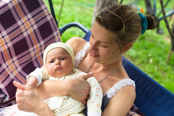 Anne ve bebek bahçede yaz bir salıncak üzerinde durmak. Kalıcı makyaj, doğal ve tedavi olmadan anne. Bebek üç aylık küçük bir kız gibi. Çocuk yawns.