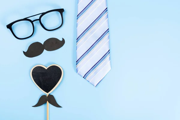 Happy Father Day background concept with blue necktie, glasses, black heart shape and black mustache on blue background with copy space for text.