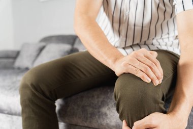 Evdeki kanepede otururken diz ağrısı çeken genç bir adam. Sağlık hizmeti tıbbi veya günlük yaşam konsepti.