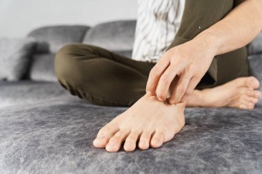 Evdeki kanepede otururken ayağını kaşıyan genç adam. Sağlık hizmeti tıbbi veya günlük yaşam konsepti.