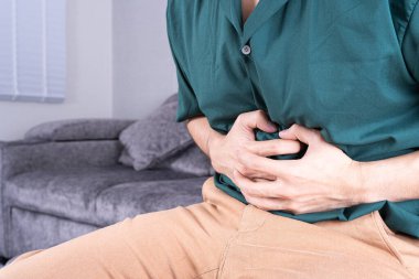 Evdeki kanepede otururken karın ağrısı çeken genç bir adam. Sağlık hizmeti tıbbi veya günlük yaşam konsepti.