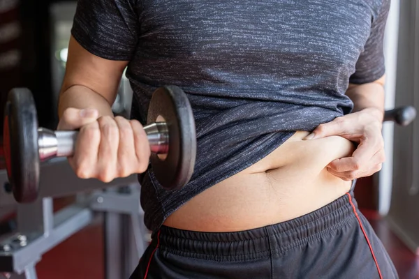 Dambıl tutan şişman bir adam ve aşırı şişman bir göbek fitness salonunda şişman bir göbek. Diyet yaşam tarzı, kilo kaybı, mide kası, sağlıklı konsept.