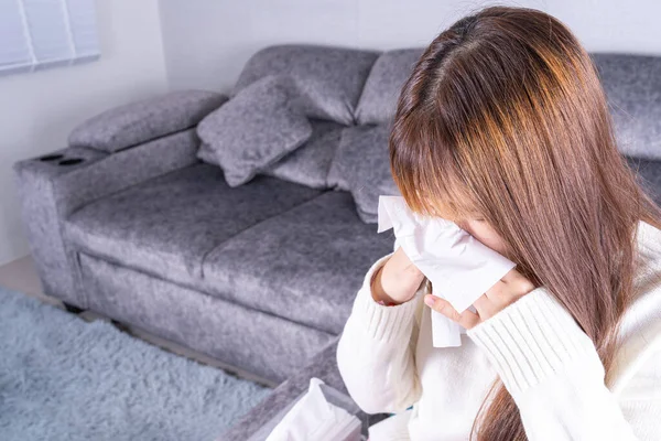 Asian beautiful woman blowing a nose to tissue while sitting on sofa at home. Healthcare medical or daily life concept.