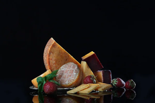 A large round piece of cheese on a black background with vintage dishes and strawberries. Copy space. Macro photo. The concept of craft cheeses. — Stock Photo, Image