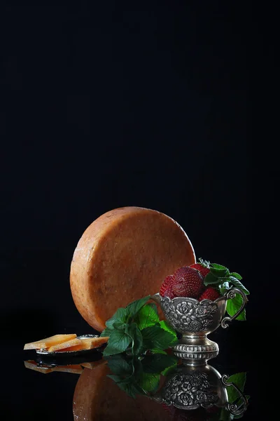 A large round piece of cheese on a black background with vintage dishes and strawberries. Copy space. Macro photo. The concept of craft cheeses. — Stock Photo, Image