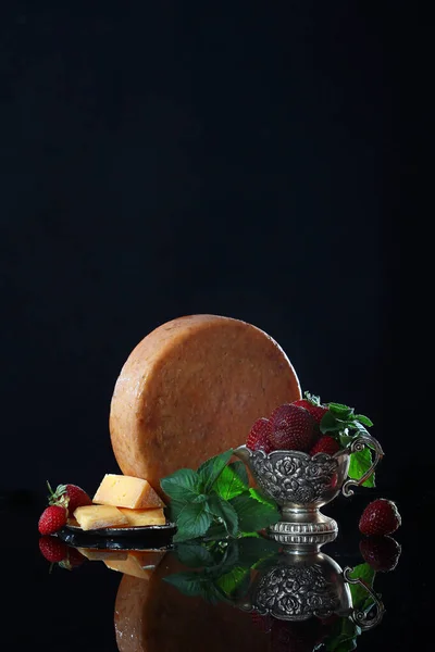 A large round piece of cheese on a black background with vintage dishes and strawberries. Copy space. Macro photo. The concept of craft cheeses. — Stock Photo, Image