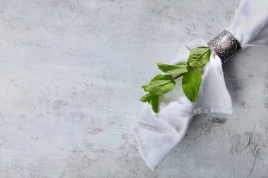 Beyaz bir pamuk peçete antika bir metal halkayla bağlanır. Hafif desenli bir arka planda dekoratif eşyalar. Bir tutam nane servisi süslüyor. Boşluğu kopyala. Üst görünüm.