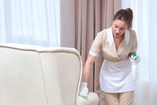 Cleaning service at the hotel. A girl in a uniform wipes upholstered furniture. Plastic bottle and napkin in hand. The concept of the hotel business. Copy space.