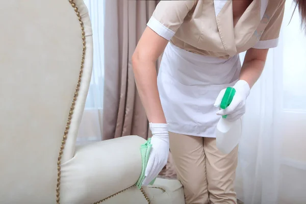 Cleaning service at the hotel. A girl in a uniform wipes upholstered furniture. Unrecognizable photo.Plastic bottle and napkin in hand. The concept of the hotel business. Copy space.