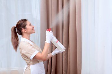 Üniformalı bir hizmetçi bir otel odasında plastik bir şişeden oda spreyi sıkar. Beyaz pamuk eldivenli eller. Uzayı kopyala Otel işi kavramı..