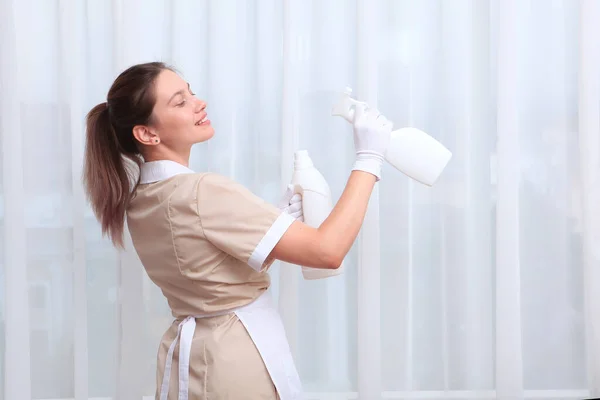 制服を着た若い家政婦が白いペットボトルを手に持っている 白い綿の手袋で手 窓の背景の写真 ホテルビジネスのコンセプト スペースのコピー — ストック写真